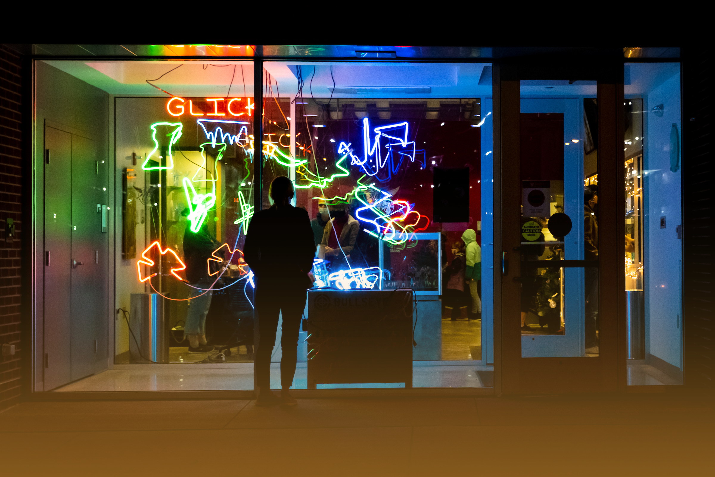 Student standing with his back facing the camera, looking through the window into the Glick Center at neon signs.